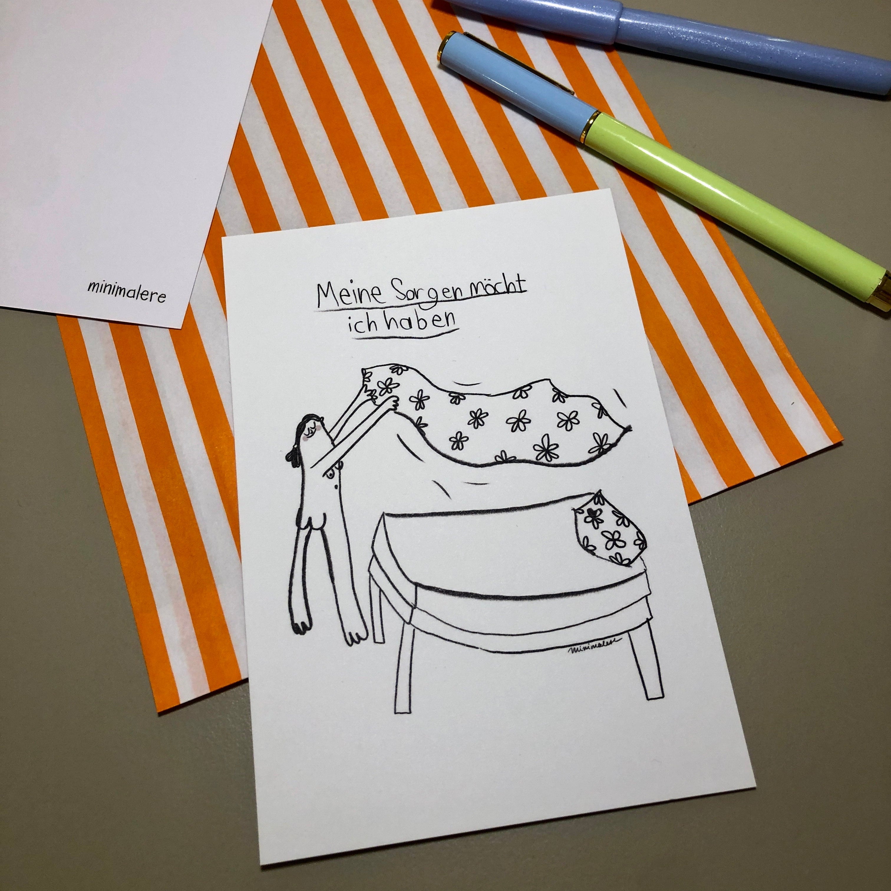 Eine Grußkarte mit der Aufschrift "Meine Sorgen möcht ich haben." liegt auf einer orange-weiß gestreiften Papiertüte, daneben liegen zwei Stifte.