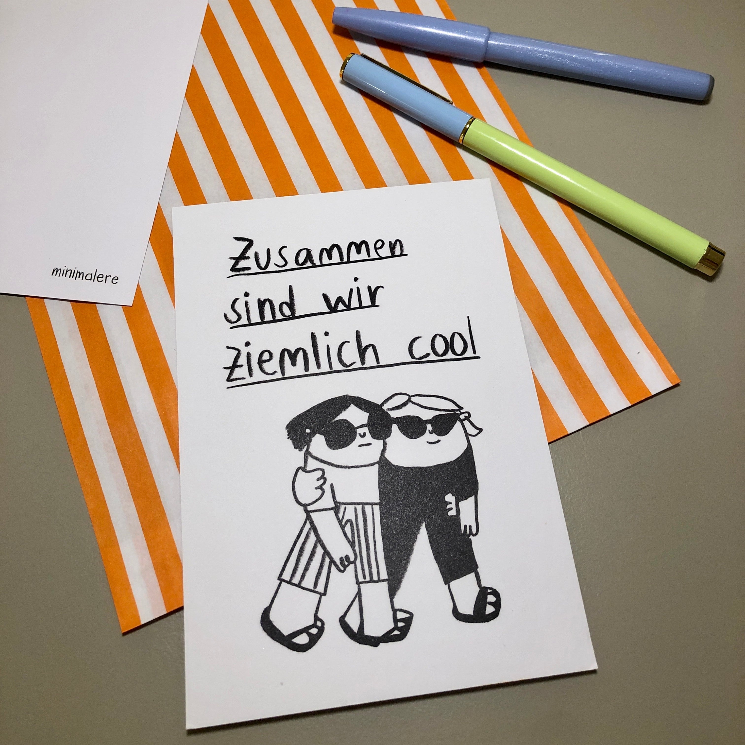 Eine Grußkarte mit der Aufschrift "Zusammen sind wir ziemlich cool." liegt auf einer orange-weiß gestreiften Papiertüte, daneben liegen zwei Stifte.