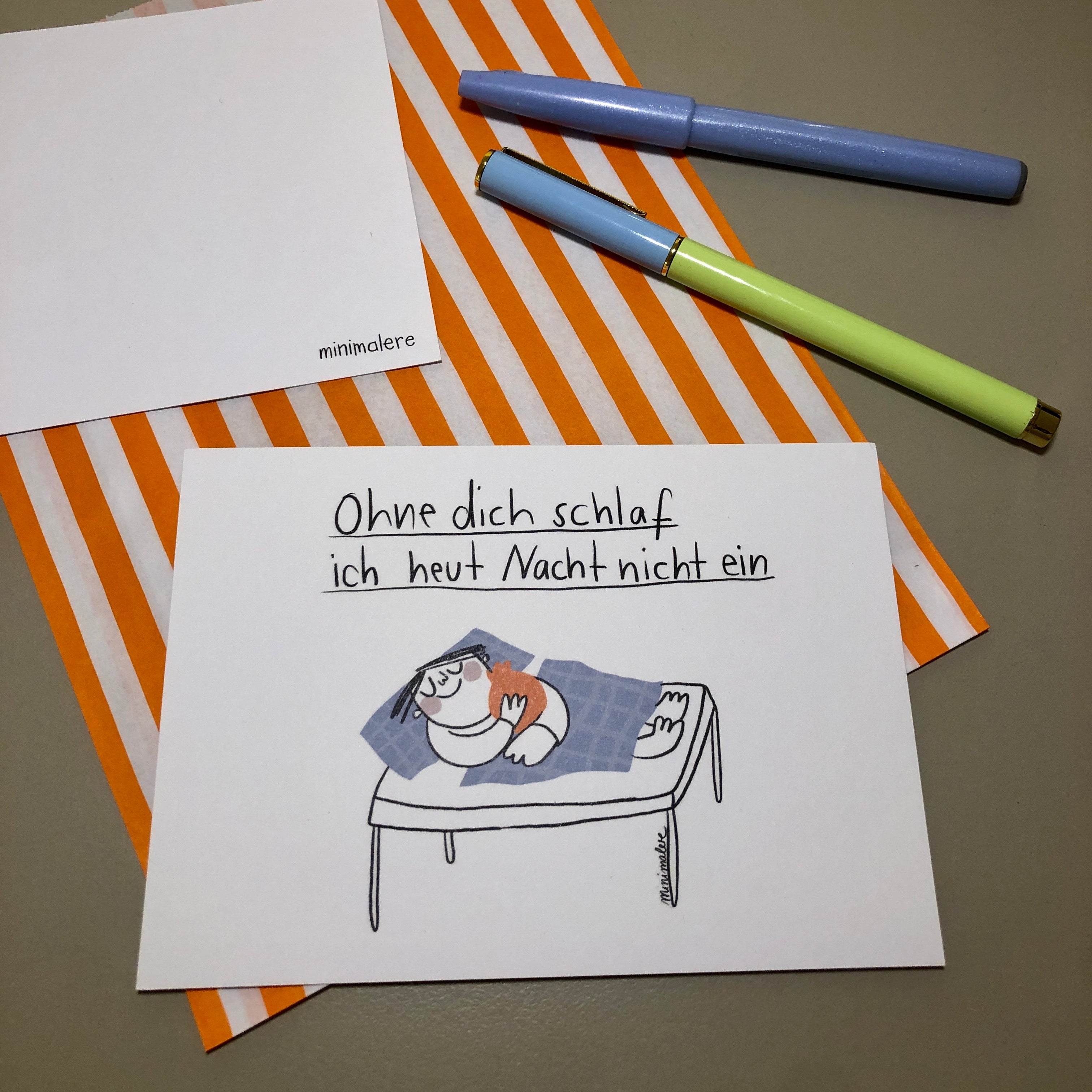 Eine Grußkarte mit der Aufschrift "Ohne dich schlaf ich heut Nacht nicht ein" liegt auf einer orange-weiß gestreiften Papiertüte, daneben liegen zwei Stifte.