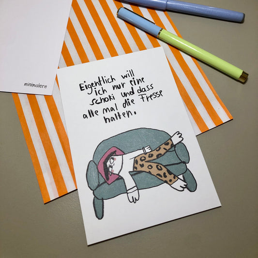 Eine Grußkarte mit der Aufschrift "Eigentlich will ich nur eine Schoko und dass alle mal die Fresse halten." liegt auf einer orange-weiß gestreiften Papiertüte, daneben liegen zwei Stifte.