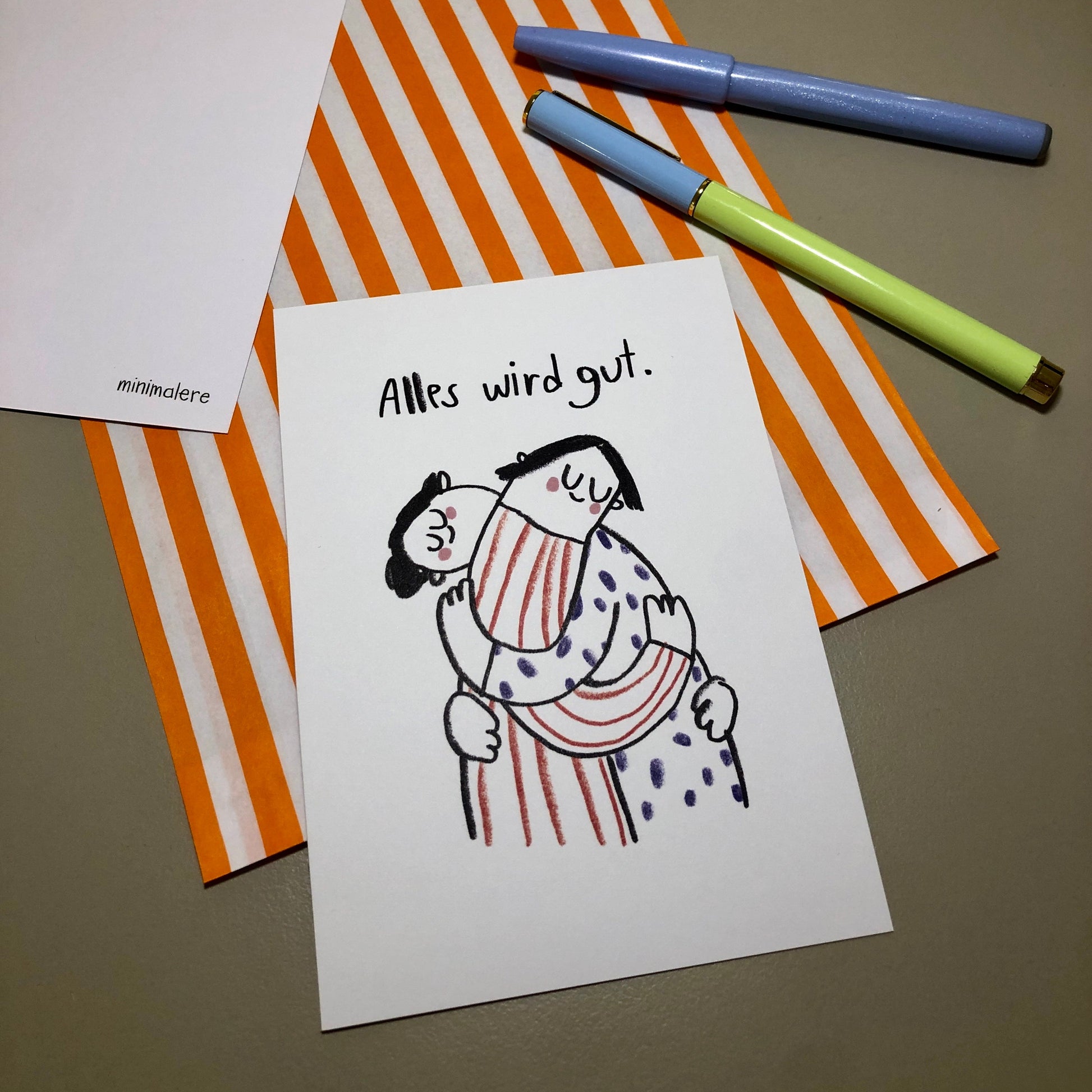 Eine Grußkarte mit der Aufschrift "Alles wird gut." liegt auf einer orange-weiß gestreiften Papiertüte, daneben liegen zwei Stifte.