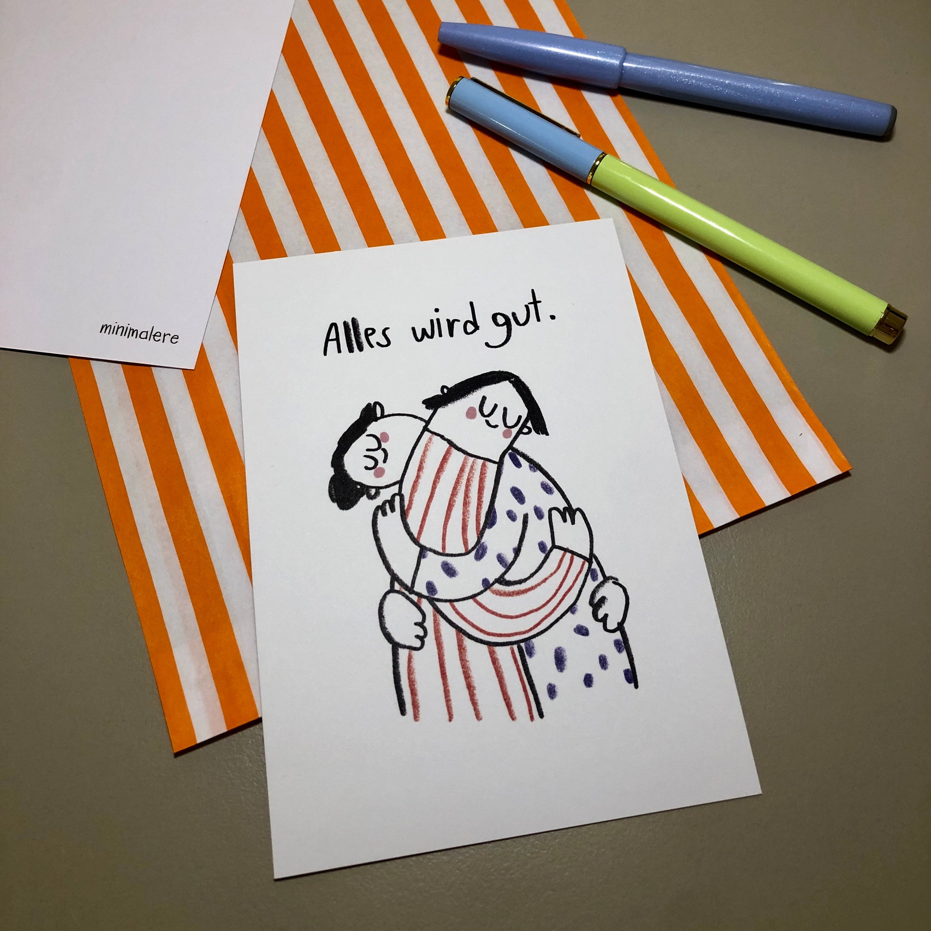 Eine Grußkarte mit der Aufschrift "Alles wird gut." liegt auf einer orange-weiß gestreiften Papiertüte, daneben liegen zwei Stifte.