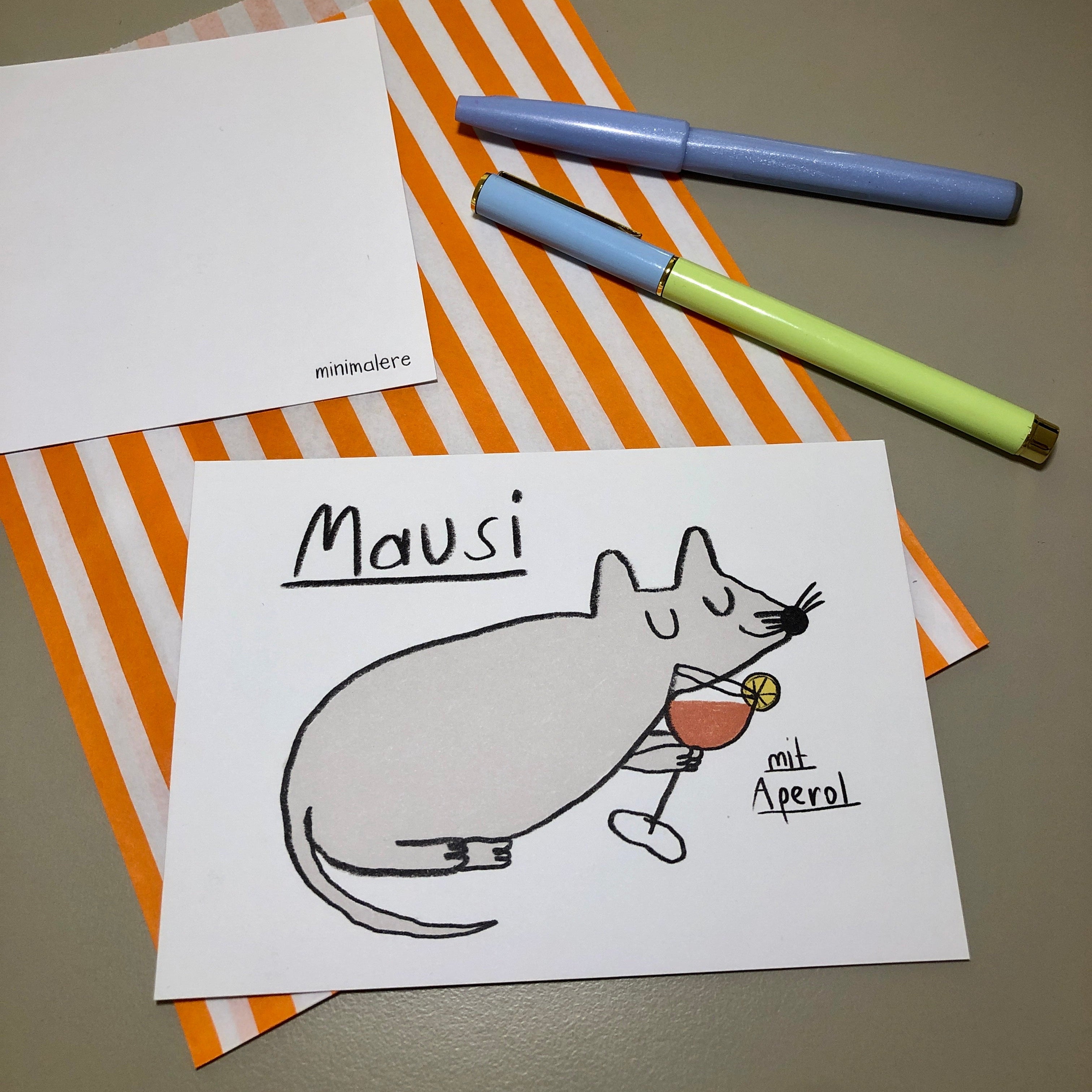 Eine Grußkarte mit der Aufschrift "Mausi mit Aperol." liegt auf einer orange-weiß gestreiften Papiertüte, daneben liegen zwei Stifte.