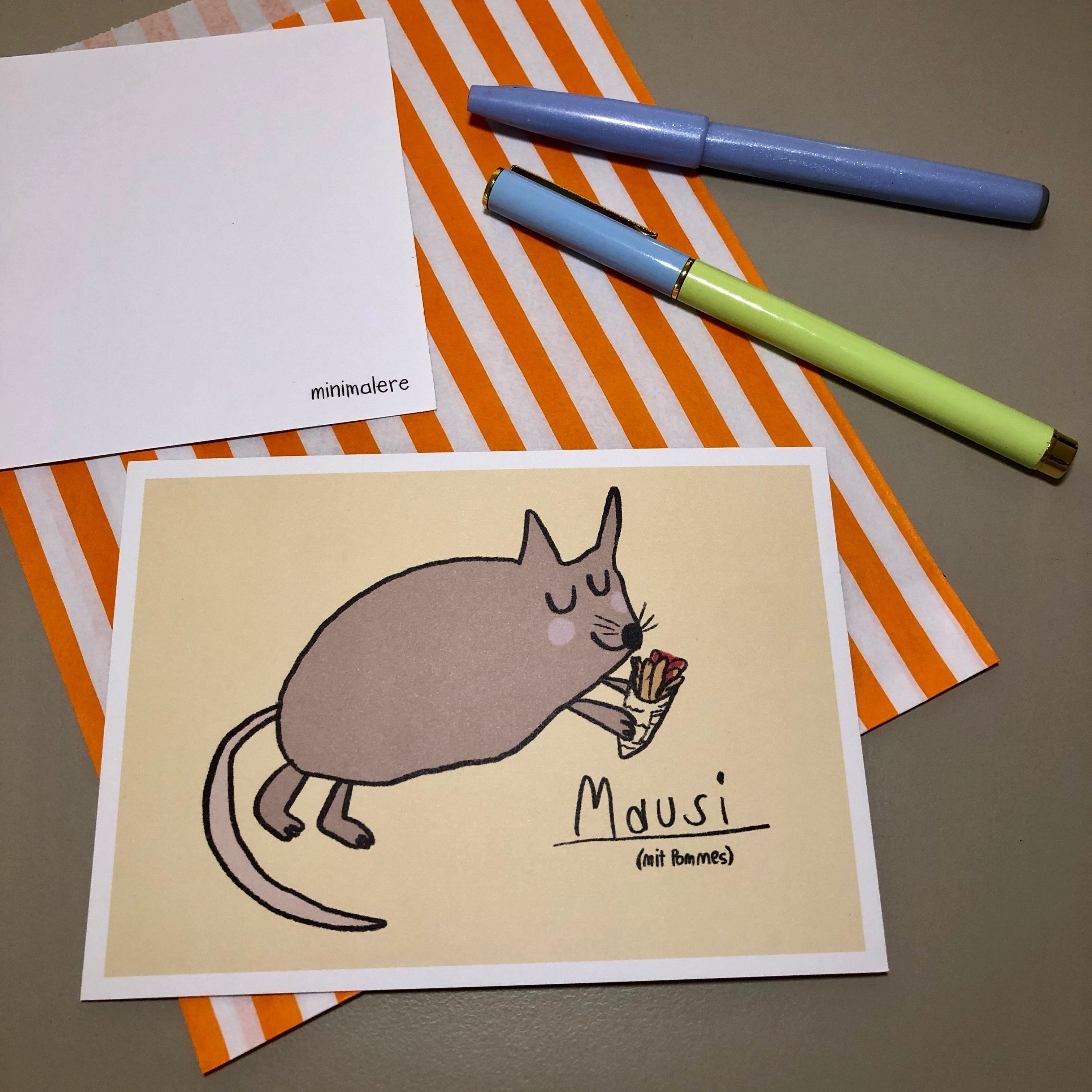 Eine Grußkarte mit der Aufschrift "Mausi (mit Pommes)." liegt auf einer orange-weiß gestreiften Papiertüte, daneben liegen zwei Stifte.